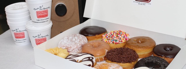 An open box of a dozen assorted donuts next to Donut Station coffee cups